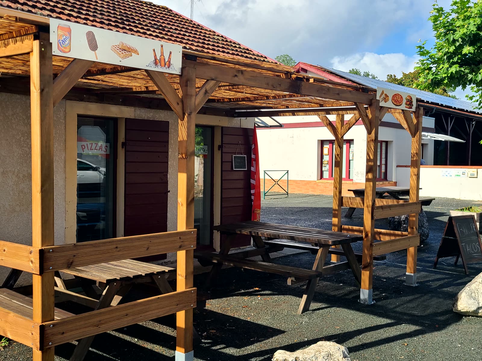 La pizzeria Pizza Dello à Taller : volets bordeaux, terrasse sous une pergola en bois avec tables de pique-nique, ardoise « Menu tacos » devant.